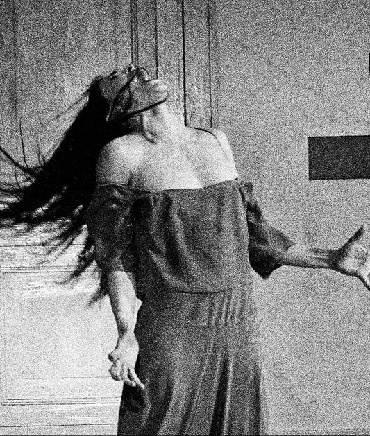 Black and white photograph of a woman in an off-the-shoulder dress, leaning back with her arms outstretched, standing in front of a paneled door and textured wall.