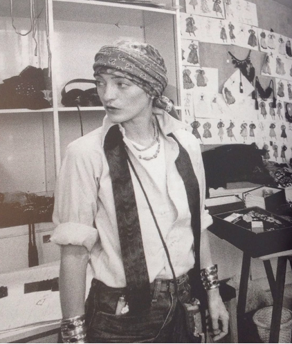 A black and white photograph of a person wearing a patterned headscarf, a light-colored shirt with a dark fuzzy collar, layered necklaces, and a crossbody bag, standing in what appears to be a fashion studio or workshop with shelves and sketches on the wall.