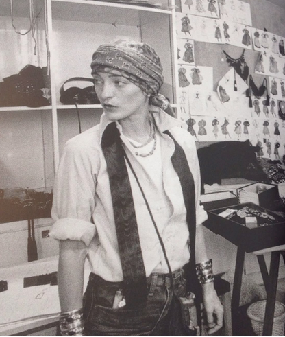 A black and white photograph of a person wearing a patterned headscarf, a light-colored shirt with a dark fuzzy collar, layered necklaces, and a crossbody bag, standing in what appears to be a fashion studio or workshop with shelves and sketches on the wall.