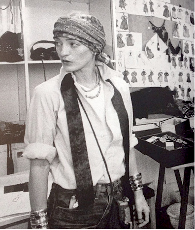 A black and white photograph of a person wearing a patterned headscarf, a light-colored shirt with rolled sleeves, a dark vest, and jeans, standing in what appears to be a fashion studio or workshop with shelves and sketches in the background.
