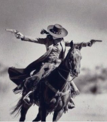 Black and white western photograph of a rider on a horse, holding two revolvers, with a wide-brimmed hat and flowing coat against a blurred outdoor background.